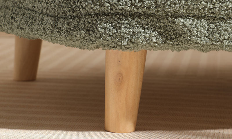 Close-up of a wooden leg supporting a textured green fabric piece of furniture on a beige carpeted floor.