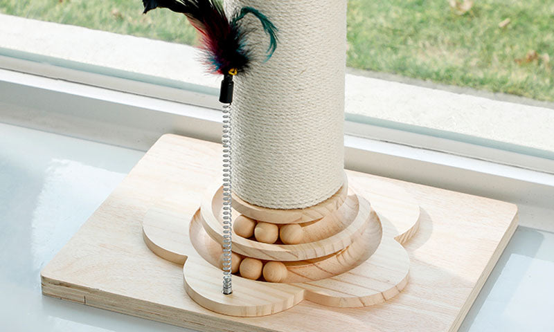 Close-up of a wooden cat scratching post base with balls and a feather toy by a window sill with grass outside